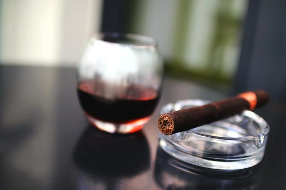 How to Smoke a Cigar — unbranded cigar resting on a glass ashtray beside a glass of red wine on a dark table